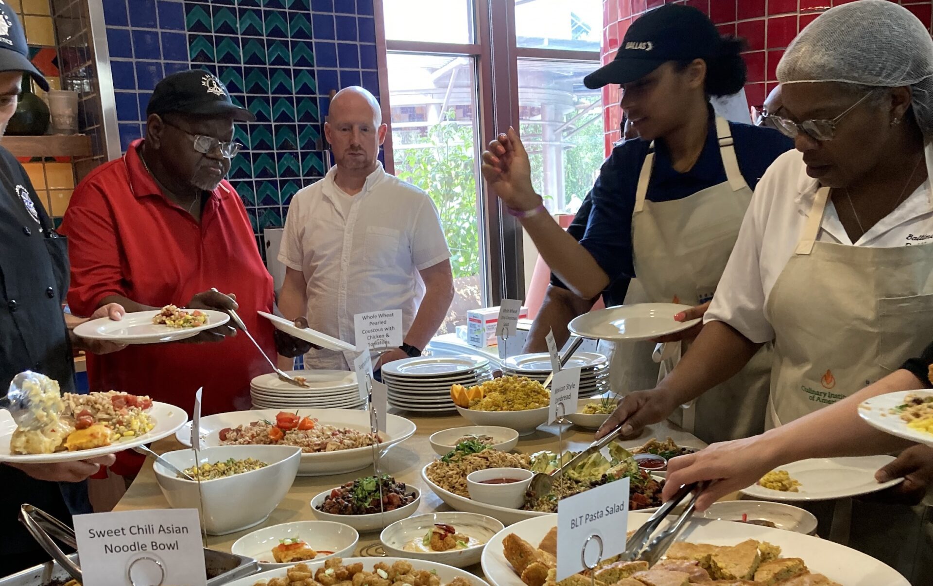 Andrew Urbanetti joins a group of people gathered around serving platters with foods and name plates, including recipes titled “Sweet Chili Asian Noodle Bowl’ and “BLT Pasta Salad.”